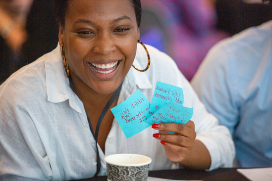 Woman sitting at a table smiling, and holding up three written-on sticky notes in front of her