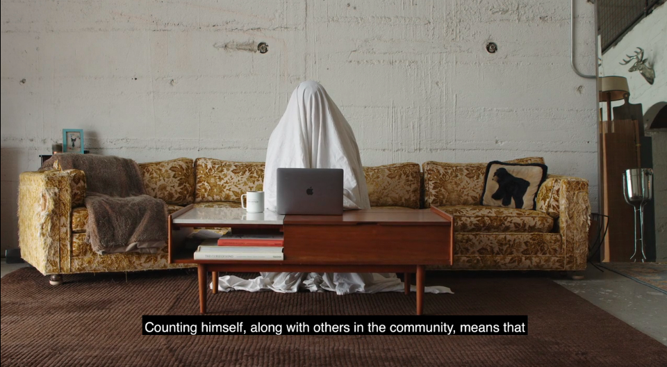 Person wearing a sheet over their head as a ghost costume, sitting on the sofa typing at a laptop