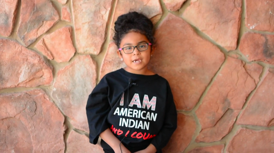 Little girl standing in front of a stone wall, wearing a black shirt that reads I am American Indian and I count